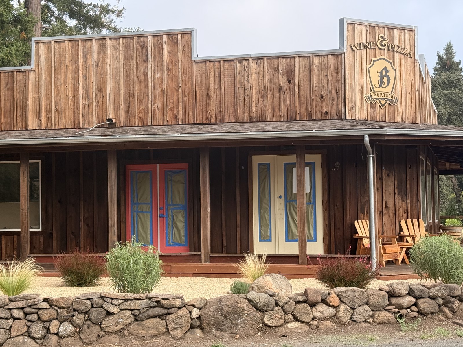 The timber facade of the Burtech building with a red door and a cream door, both masked with painter's tape, and Adirondack chairs on the porch