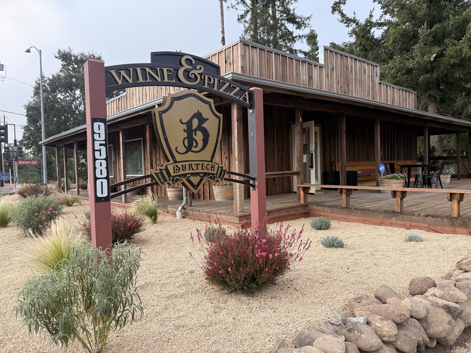 A wooden arch sign reading WINE & PIZZA above a gold Burtech shield crest, in front of a timber building at 9580 Sonoma Highway