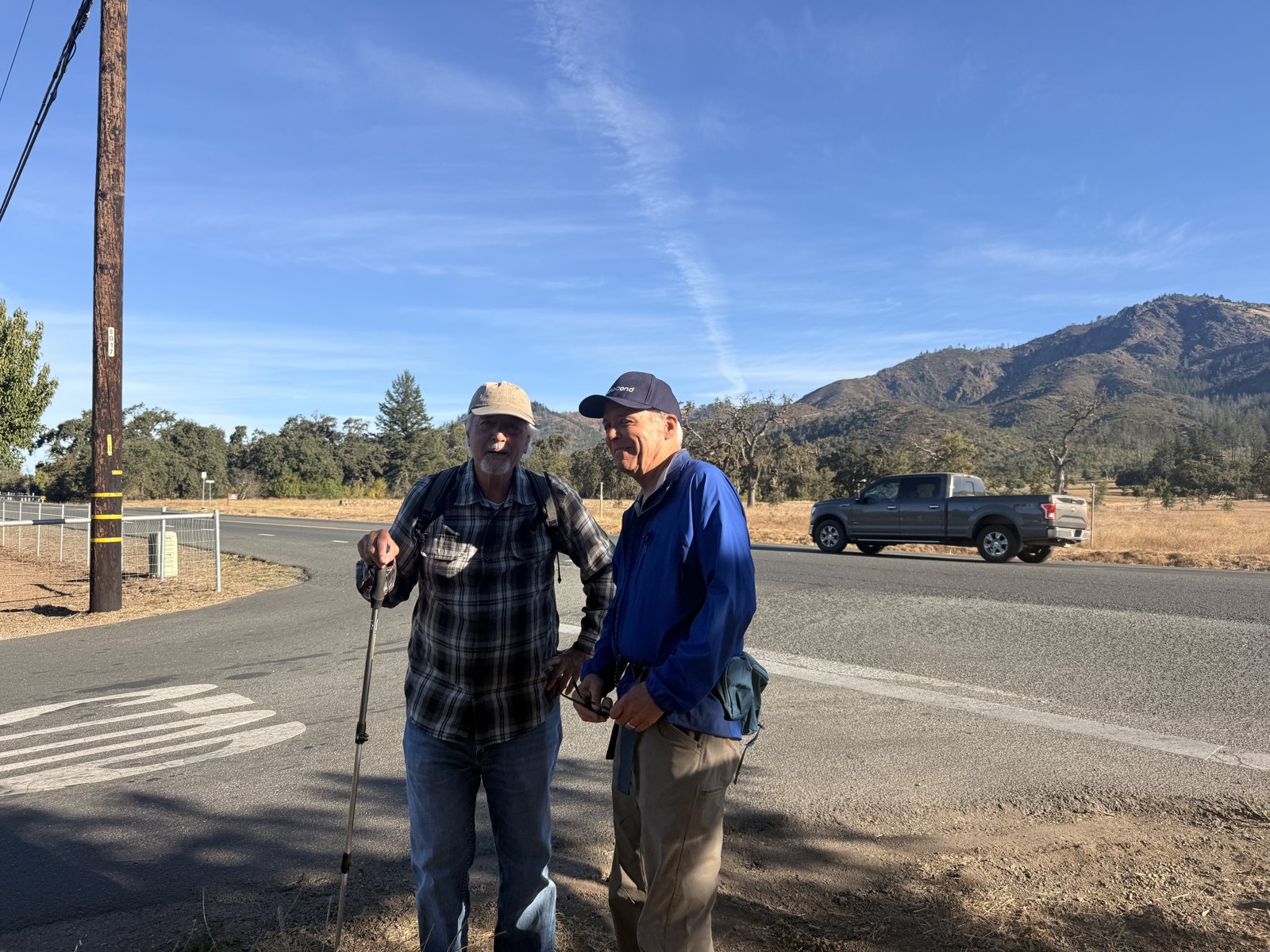 Marty Behr and Mark Campbell standing at a crosswalk on Highway 12 in Kenwood, with Hood Mountain behind them and a pickup truck passing