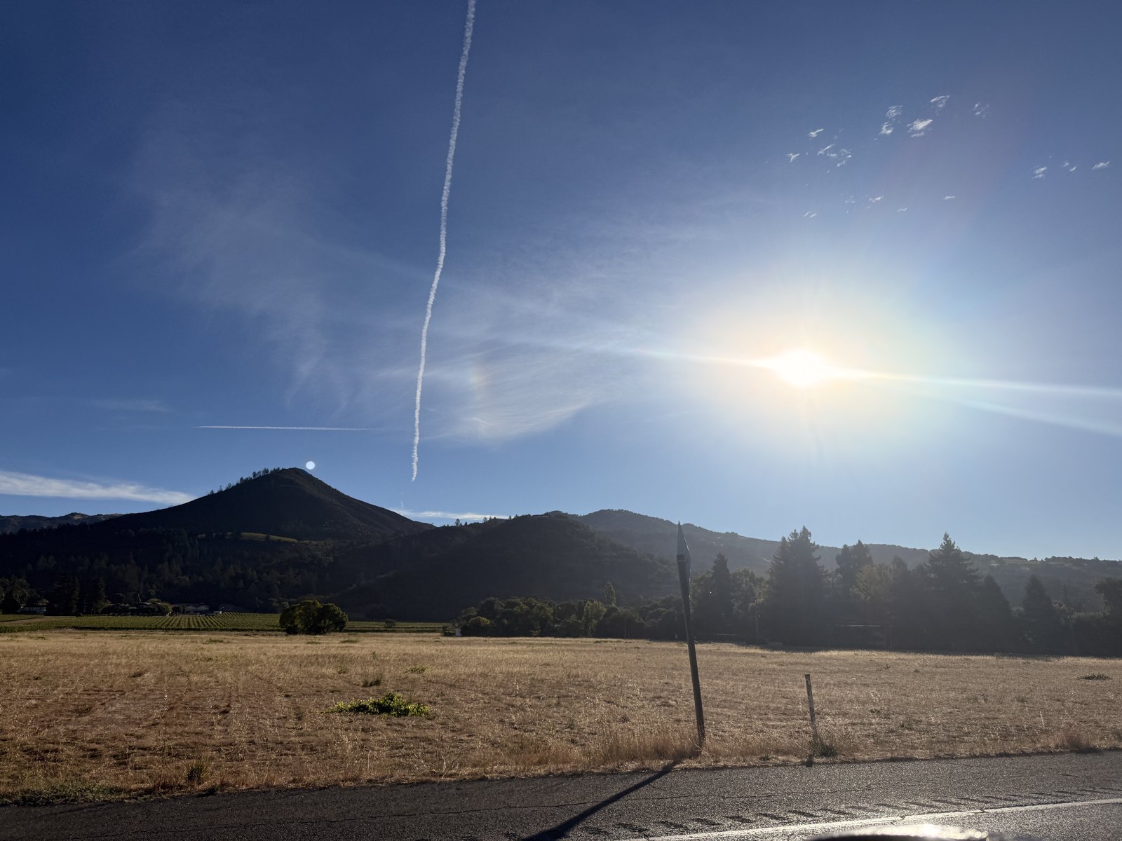 Late afternoon sun low over Hood Mountain with a bright patch of light beside it, a contrail overhead and the moon above the ridge